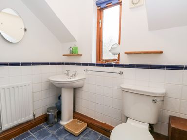 A bathroom with a pedestal sink, toilet, small window, round wall mirror, and shelves at Friars Fold in Newby Bridge