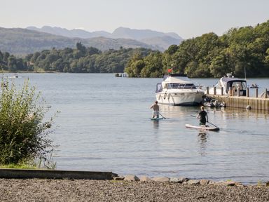Two people paddleboarding on a lake near boats and a dock with trees and hills in the background at Friars Fold in Newby Bridge
