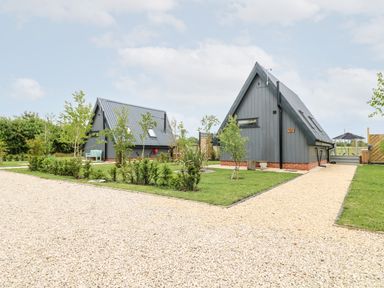 Two small gray A-frame houses with metal roofs and small trees around on a gravel path at Buttercup Lodge in Misterton