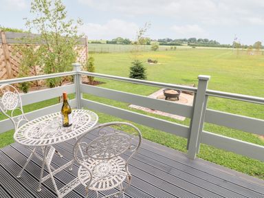 A patio with a white metal table and two chairs with a bottle on the table and a grassy field with a fire pit in the background at Buttercup Lodge in Misterton