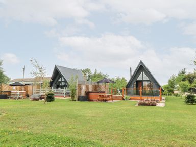 Two small A-frame cabins with fenced patios and outdoor seating on a grassy lawn at Buttercup Lodge in Misterton
