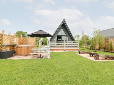 A garden with a picnic table and umbrella a wooden hot tub a fire pit with logs and a triangular cabin at Buttercup Lodge in Misterton