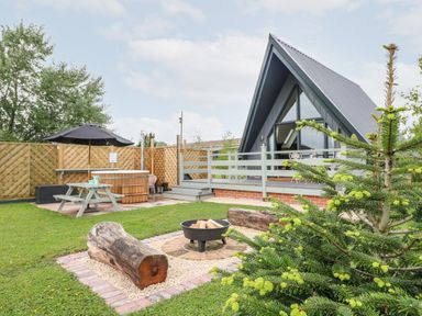 An outdoor area with a wooden hot tub table fire pit and log seating near a cabin with large windows at Buttercup Lodge in Misterton