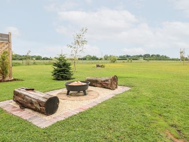 An outdoor fire pit with two large wooden logs as seating on a grass lawn at Buttercup Lodge in Misterton