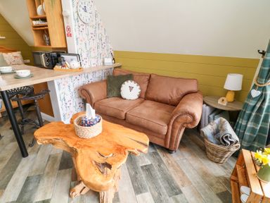 A living area with a brown sofa, a wooden coffee table, a small side table with a lamp, a basket with blankets, and a kitchen counter with chairs at Buttercup Lodge in Misterton