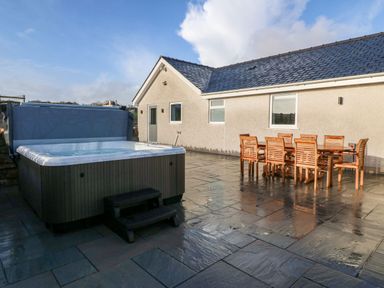 A patio with a hot tub and wooden dining table with chairs at Yr Hen Feudy in Moelfre