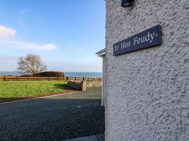 View of a stone house exterior wall with a sign Yr Hen Feudy and a fenced grassy yard with a tree and ocean in background at Yr Hen Feudy in Moelfre
