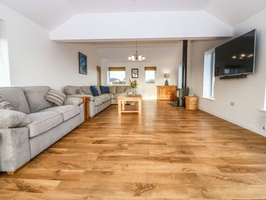 A living room with gray sofas a coffee table a wood stove and a wall-mounted television at Yr Hen Feudy in Moelfre