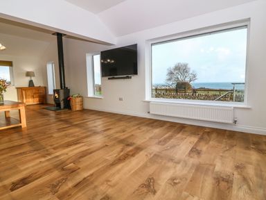 A living room with a large window overlooking the sea a wood stove and a wall-mounted television at Yr Hen Feudy in Moelfre