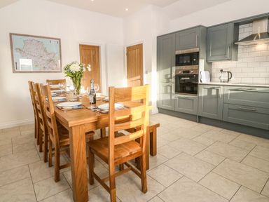 A kitchen and dining area with a wooden dining table set with plates and glasses and grey cabinets with built-in oven at Yr Hen Feudy Moelfre