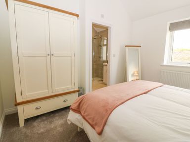 A bedroom with a bed covered with a pink blanket a cream wardrobe a full length mirror and an en suite bathroom at Yr Hen Feudy in Moelfre