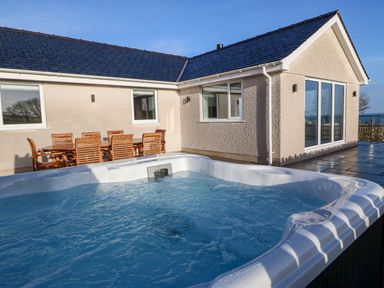 An outdoor patio with a hot tub and wooden dining table with chairs next to a house at Yr Hen Feudy in Moelfre