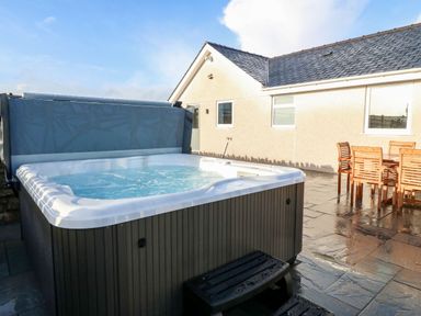 An outdoor hot tub with water and a wooden dining table with chairs on a stone patio at Yr Hen Feudy in Moelfre