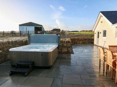 An outdoor patio area with a hot tub wooden chairs and table stone wall and farm buildings in the background at Yr Hen Feudy in Moelfre