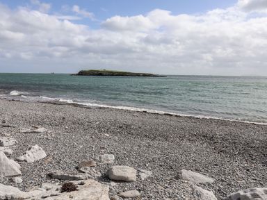 A pebble beach with rocks near the shore and an island in the sea under a cloudy sky at Yr Hen Feudy in Moelfre