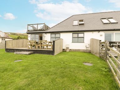 An outdoor patio with wooden table and benches adjacent to a house with fenced grass yard at Bwthyn Awel in Church Bay near Cemaes Bay