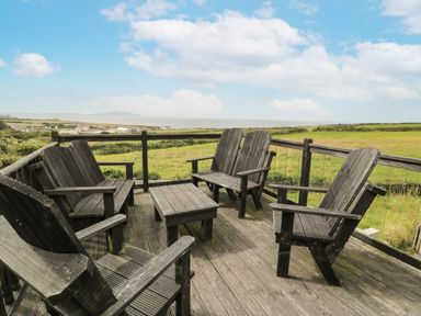 A wooden deck with five wooden chairs and a small table overlooking a field at Bwthyn Awel in Church Bay near Cemaes Bay