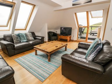 A living room with two black leather sofas a wooden coffee table a patterned blue rug a television on a wooden stand and large windows at Bwthyn Awel in Church Bay near Cemaes Bay