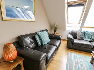 A living room with two black leather sofas blue cushions a wooden side table with a lamp and a painting on the wall at Bwthyn Awel in Church Bay near Cemaes Bay