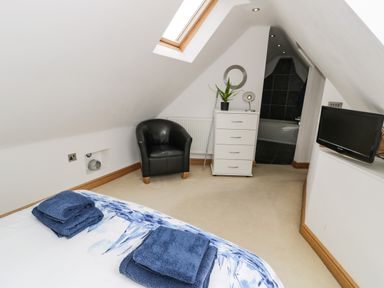 A bedroom with a bed, blue towels on the bed, a black armchair, a white chest of drawers with a plant and a mirror above, a small TV, and an open door to a bathroom at Bwthyn Awel Church Bay near Cemaes Bay