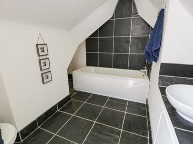 A bathroom with a white bathtub black tiled floor and walls a white sink and blue towels at Bwthyn Awel in Church Bay near Cemaes Bay