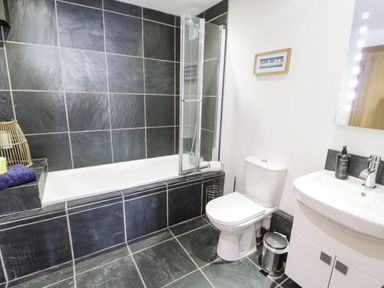 A bathroom with a bathtub surrounded by black tiles a white toilet and a white sink with a mirror at Bwthyn Awel in Church Bay near Cemaes Bay