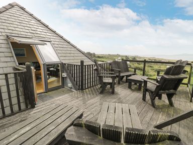 An outdoor wooden deck with chairs and a small table next to a pitched roof house overlooking a rural landscape at Bwthyn Awel in Church Bay near Cemaes Bay