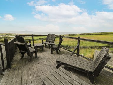 A wooden deck with chairs and a table overlooking green fields and distant hills at Bwthyn Awel in Church Bay near Cemaes Bay
