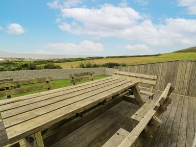 An outdoor wooden picnic table with benches on a deck overlooking fields at Bwthyn Awel in Church Bay near Cemaes Bay