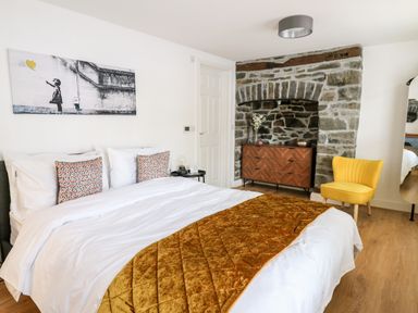 A bedroom with a bed white bedding and a gold blanket a wooden dresser set in a stone alcove and a yellow chair at Bridgend Cottages in Howey near Llandrindod Wells