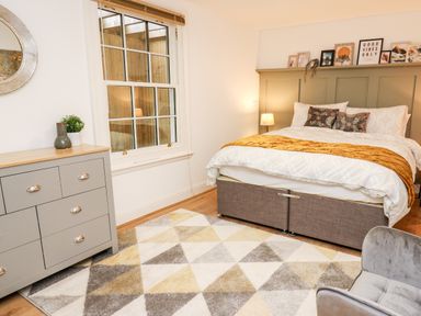 A bedroom with a bed framed pictures a chest of drawers a geometric rug and a grey chair at Bridgend Cottages in Howey near Llandrindod Wells