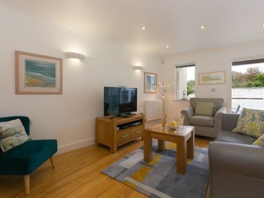 A living room with gray and green chairs a wooden table with drinks and snacks and a TV on a wooden stand at The Waves in St Ives