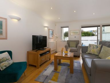 A living room with a television on a wooden stand a coffee table with snacks and drinks a grey armchair a grey sofa and a green chair at The Waves in St Ives