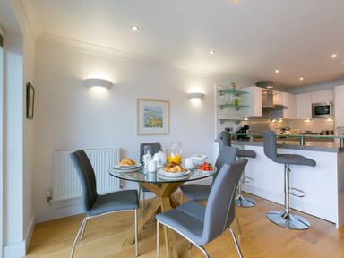 A dining area with a glass table set with croissants and orange juice and grey chairs next to a kitchen with bar stools at The Waves in St Ives