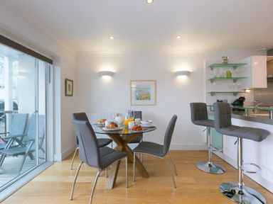 A dining area with a glass table, six gray chairs, breakfast items on the table, two bar stools by a kitchen counter and sliding glass doors leading to an outdoor seating area at The Waves in St Ives