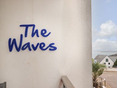 A white wall with the words The Waves in blue with a railing and a view of a building and the sea at The Waves in St Ives