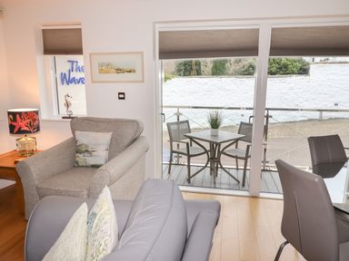A living room with armchair sofa glass dining table and chairs next to a balcony with outdoor table and chairs at The Waves in St Ives