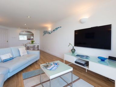 A living room with a light blue sofa a coffee table magazines a wall-mounted TV and a dining table in view at Turquarze in Carbis Bay