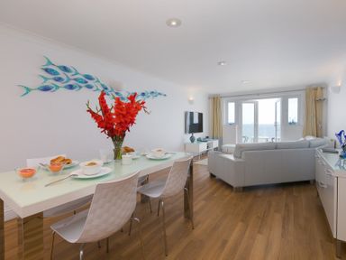 A dining area with a table set for breakfast and a living room with sofas facing a television and glass doors overlooking the sea at Turquarze in Carbis Bay