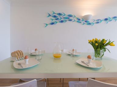 A dining table set with plates holding boiled eggs and spoons a glass jug of orange juice a toast rack with sliced bread and a vase of yellow tulips at Turquarze Carbis Bay