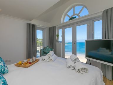 A bedroom with a bed, rolled towels, a breakfast tray, an armchair, a TV and a large window with a sea view at Turquarze in Carbis Bay