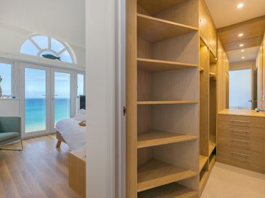 A bedroom with large windows overlooking the sea and an adjoining walk in closet with wooden shelves and drawers at Turquarze in Carbis Bay