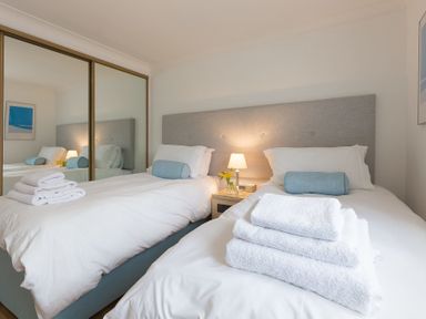 A bedroom with two single beds white linens blue bolsters towels on beds a bedside table lamp and flowers at Turquarze in Carbis Bay