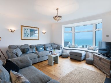 A living room with grey sofas a small wooden table large windows with a sea view and a wooden TV stand at Whitsunday House in Carbis Bay