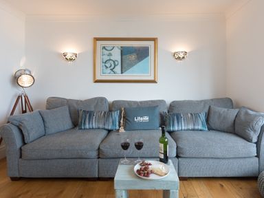 A gray sofa with cushions a small table with wine glasses a bottle cheese and grapes in a living room at Whitsunday House in Carbis Bay