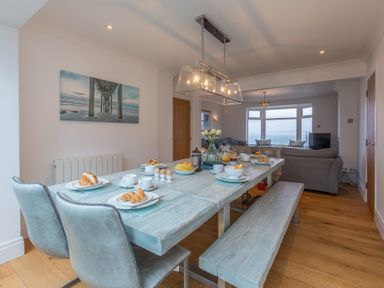 A dining area with a wooden table set for breakfast adjacent to a living room with a sofa and large windows showing a sea view at Whitsunday House in Carbis Bay