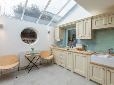 A kitchen with cream cabinets a wooden countertop a farmhouse sink two wooden chairs a small table with flowers and a circular window at Whitsunday House in Carbis Bay