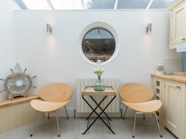 A sitting area with two wooden chairs a small table with a vase of flowers a round window and kitchen cabinets at Whitsunday House in Carbis Bay