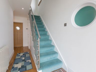 A hallway with blue carpeted stairs a wooden door with round window and a rug on wooden floor at Whitsunday House in Carbis Bay