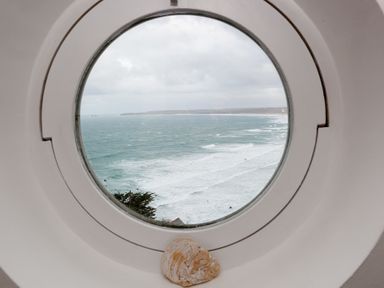 A round window looking out onto a beach and ocean with waves and a cloudy sky at Whitsunday House in Carbis Bay